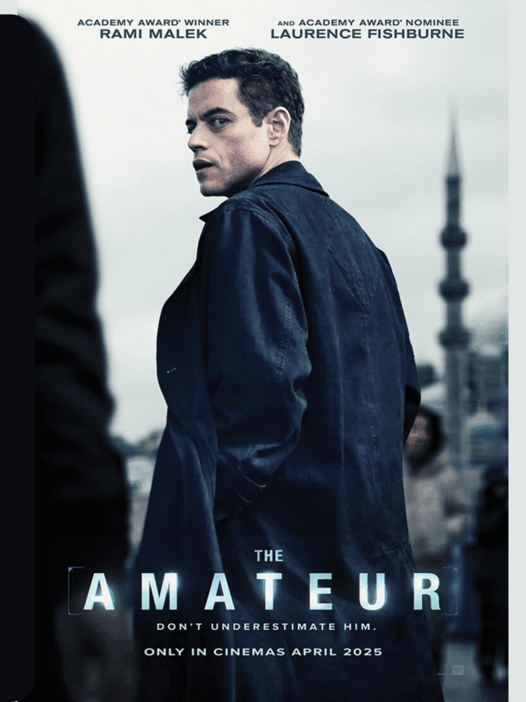 The Amateur movie poster showing Rami Malek in a dark coat looking back with a cityscape and tower blurred behind him.