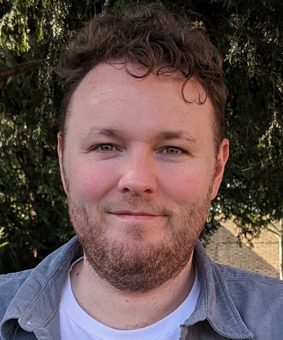 Close-up portrait of Brendan Duffy outdoors with leafy trees and brick building in background