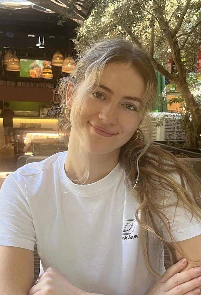 Young woman with long wavy hair smiling indoors near a tree and hanging lights.