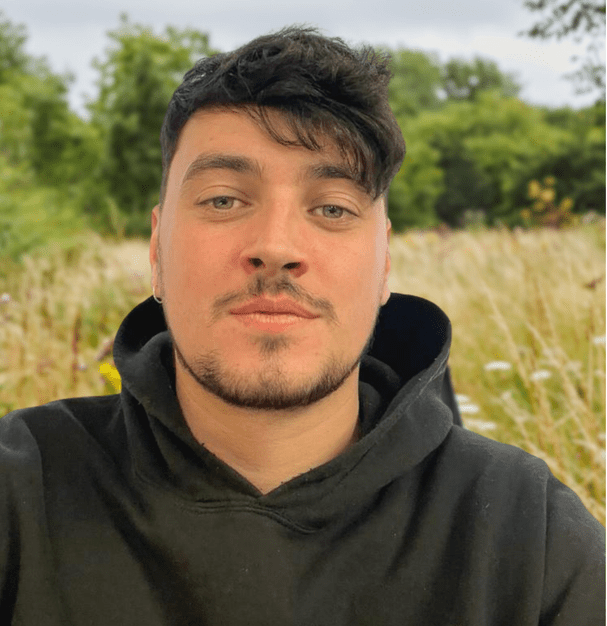 Close-up of a man with dark hair and beard wearing a black hoodie outdoors with greenery behind