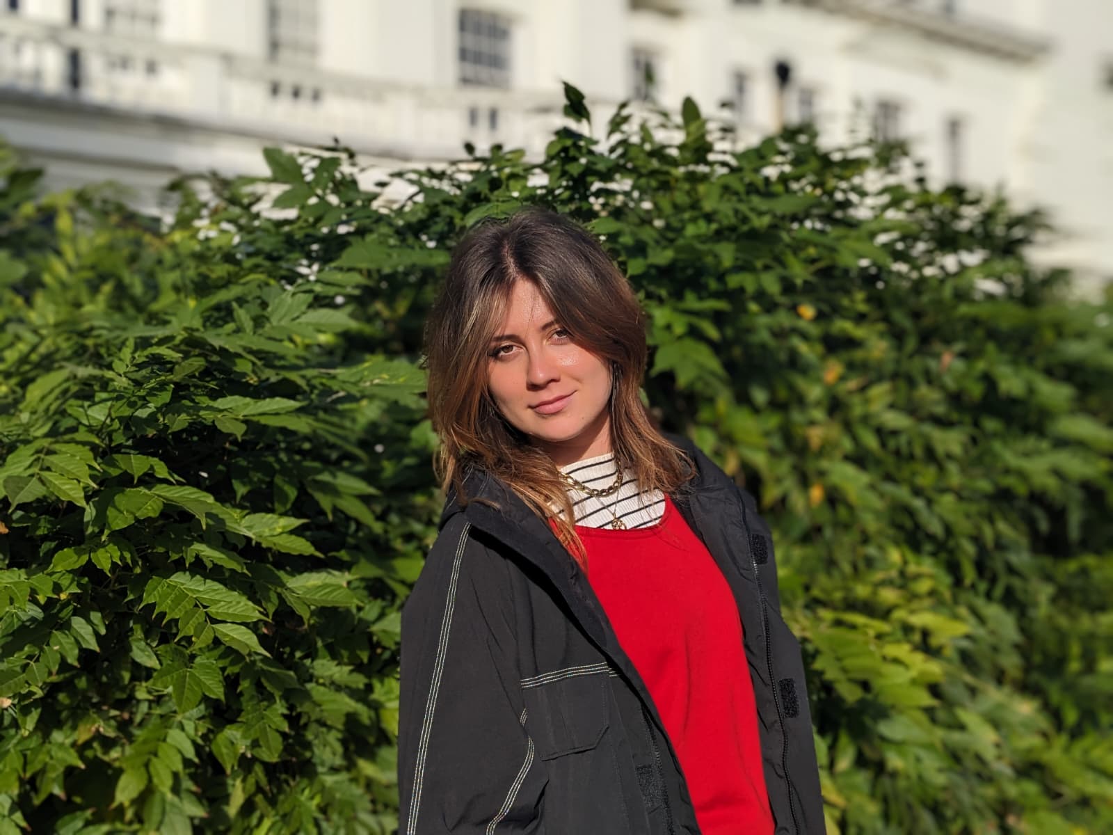 Young woman in red top and black jacket standing outdoors in front of green bushes